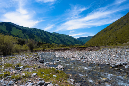 Scenic mountain river flowing through a rocky valley with green slopes and a bright blue sky with wispy clouds.