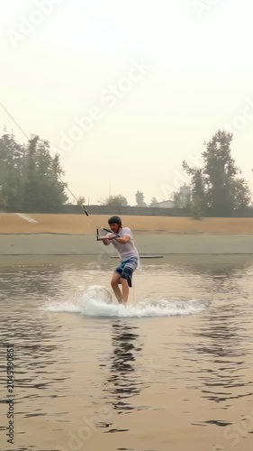 Vertical video: Cable towing rider from water onto wakeboard, carving past box on lake in helmet