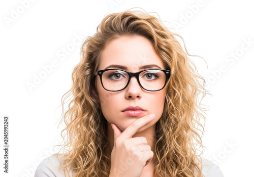Thoughtful young woman with curly blonde hair wearing glasses, hand on chin, contemplating, serious expression, intelligent look.