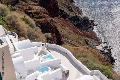 Fotografie A high-angle horizontal view of a luxury white boutique hotel with private outdoor jacuzzis, built on the steep volcanic cliff overlooking the sea in Oia, Santorini, Greece