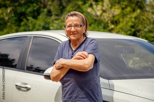 Old woman driving a car in the countryside, road trip concept.  Portrait of an old woman driving and near a car, on a walk outside the city.