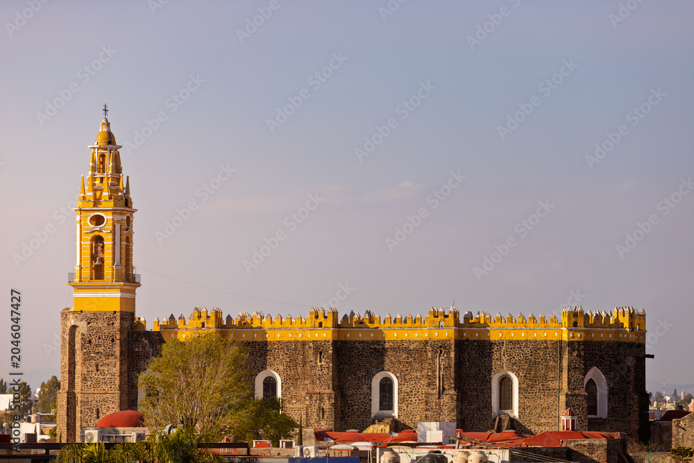 Obraz premium Exterior view of the Royal Chapel of Naturales in Cholula, featuring its distinctive yellow facade with crenellations and stone bell tower against a clear sky