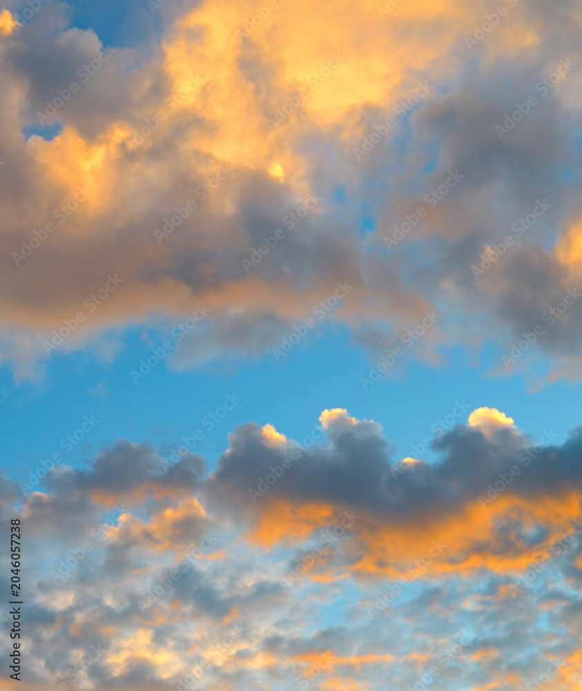 Obraz premium Premium vertical view of a beautiful dramatic sky filled with thick sunset clouds illuminated by bright orange golden hour light against a calm blue background.