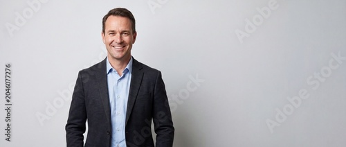 Smiling middle aged businessman in dark suit and light blue shirt standing against plain background