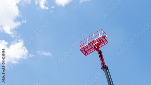 A red elevated work basket on a raised boom stands out against a clear blue sky with soft white clouds, creating a simple industrial scene with open space and bright daylight