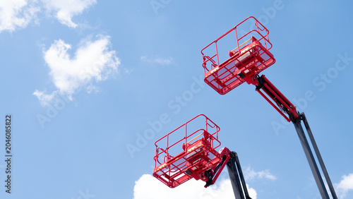 Two red aerial work platforms raised high against a clear blue sky with soft white clouds, viewed from below in an industrial maintenance setting