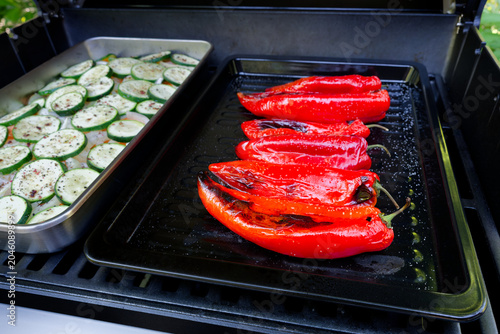 Fresh red pointed peppers and seasoned zucchini slices are cooking on a barbecue grill. A close-up of a gas grill loaded with fresh vegetables.