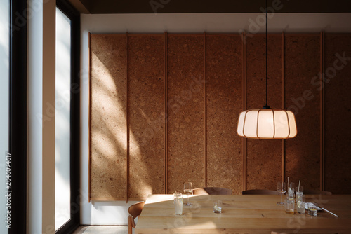An eye-level, indoor shot of a modern dining room featuring a long wooden table, a minimalist fabric pendant light, and a textured cork panel wall, bathed in soft, natural window light.