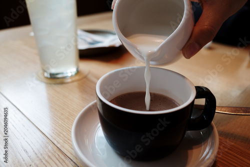 Fotografie A close-up, eye-level photograph of a hand pouring fresh milk from a small white ceramic pitcher into a dark cup of hot chocolate or coffee, served on a wooden table in a cozy cafe setting