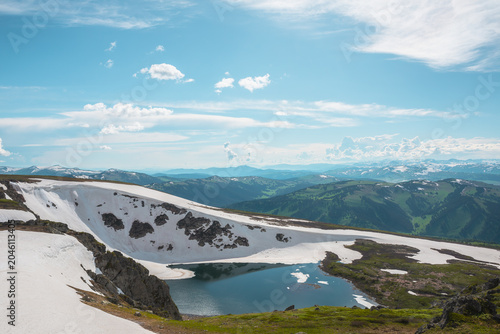 Scenic sunlit landscape with alpine lake in rocky snowy cirque near stone hill top during thaw. Ice floats in mountain lake among rocks and snows with view to forest mountain range under cloudy sky.