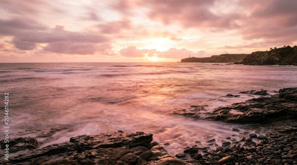 Obraz premium The sun rises over a rocky coastline, casting a warm glow on the ocean and sky. Waves gently crash against the dark, textured rocks.