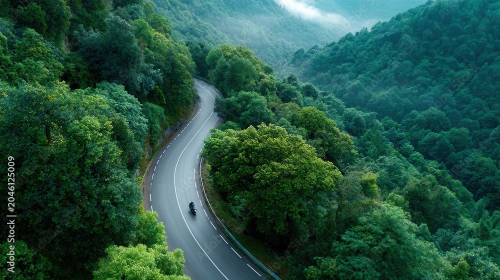 Fototapeta premium Motorcycle riding on winding mountain road through dense green forest with distant mist aerial view