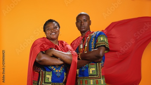 Cuadro en lienzo Ethnic married couple in superhero red capes posing on camera, feeling proud about their abilities over orange background
