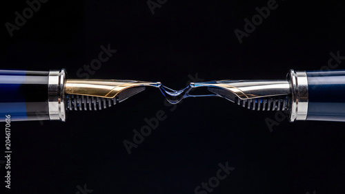 Two elegant fountain pen nibs touch, sharing a vibrant blue ink drop on a dark background.