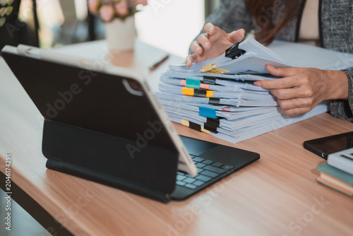Photos A close-up image of a businesswoman's hands rummaging through a large stack of documents on her desk, organizing papers and files in her office using a digital tablet