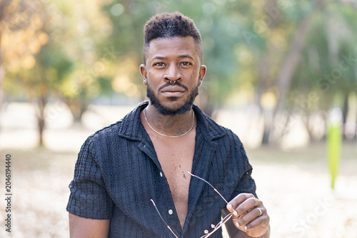 Canvas-taulu Stylish Black Man with Piercings and Beard Posing Outdoors in Soft Sunlight