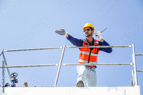Foto Male construction worker or engineer wearing safety vest and hard hat, holding a walkie talkie and pointing to direct industrial site operations on a high platform against clear blue sky background