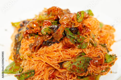 Spicy golbaengi somyeon with seasoned sea snails, fresh vegetables, and thin wheat noodles mixed in a savory Korean red pepper sauce on a white plate.