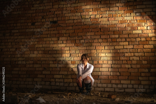 Young woman in white dress and high heels squats on uneven ground in an industrial setting with exposed brick walls and scattered debris in the background