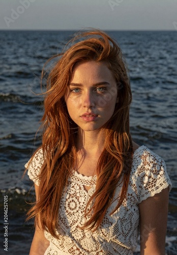 Red-haired woman in white lace dress standing by the ocean at sunset, windswept hair and serene gaze.