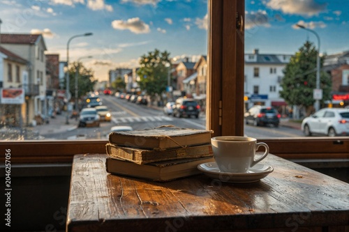 Cozy cafe window scene with stacked vintage books and a steaming coffee cup, overlooking a bustling urban street at golden hour.