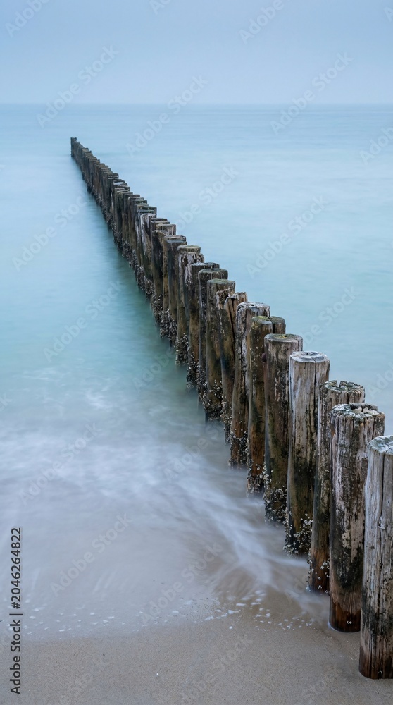 Obraz premium Dramatic perspective view of weathered wooden groyne posts leading into calm misty sea water at coastline on overcast day trip.