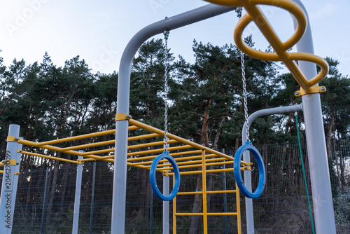 Blue gymnastic rings hanging on chains from yellow and grey calisthenics bar frame against pine forest and clear sky in outdoor public park. Concept of street workout equipment, body weight training