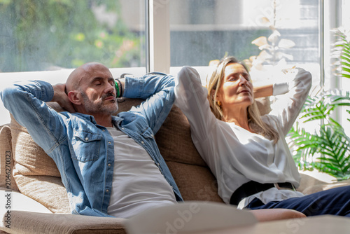 Caucasian middle-aged couple relaxing on sofa with eyes closed, hands behind heads, enjoying peaceful moment together in bright living room