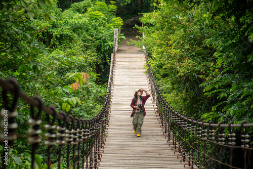 A Solo Female Explorer Walking Across A Rustic Suspension Bridge Deep In The Lush Green Rainforest