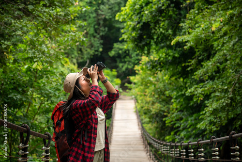 Woman Hiker With Backpack Uses Binoculars On A Wooden Suspension Bridge In A Lush Tropical Forest