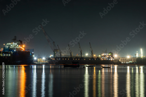 Industrial Cargo Ship Docked At A Night Port With Illuminated Cranes And Glowing Reflections On Calm Water