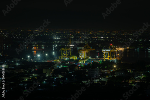Industrial Shipping Terminal Illuminated At Night By Towering Harbor Cranes Above Stacked Cargo Containers