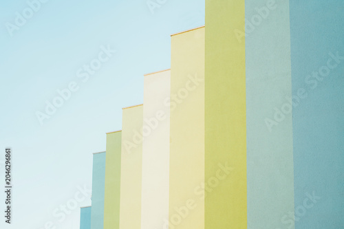 Low-angle view of a row of stepped building facades painted in soft pastel hues including blue, green, yellow and cream, set against a pale clear sky. Clean