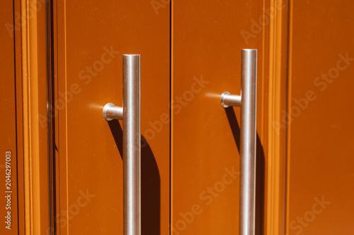 Close-up view of two vertical stainless steel tubular door pulls mounted on an orange painted double door. The symmetrical handles cast soft shadows on the