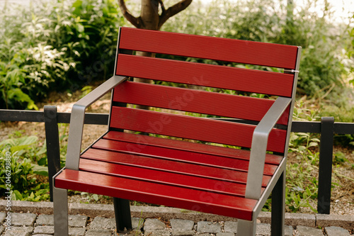 Close-up view of a red slatted wooden bench with gray metal armrests and legs positioned on a cobblestone path beside a low metal railing. Soft natural light