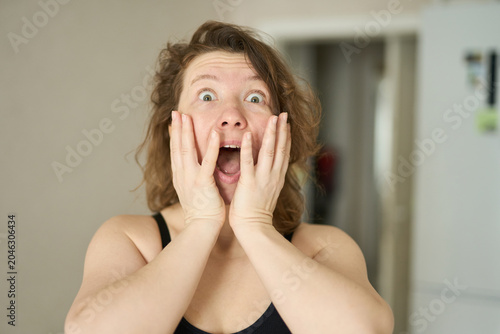 Woman With Hands On Cheeks Expressing Shock Wide Eyed Surprise In Small Home Kitchen Interior Morning Domestic Setting Casual Sleeveless Top Short Brown Hair Light Skin Standing.