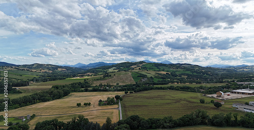 Photos Panoramica di colline coltivate e campi verdi alternati a zone boschive, con strada rurale e strutture agricole sullo sfondo, sotto cielo parzialmente nuvoloso