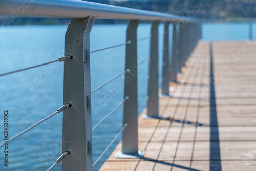 Quadro su tela Modern metal railing with cables near waters edge on a sunny day with a board walk patio stretching into the distance
