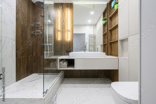 Modern bathroom with a floating white vanity, vessel sink, glass-enclosed shower, wood-toned tiles, vertical LED lighting, open shelving, and marble-effect flooring