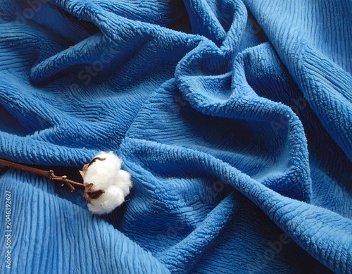 Close-up of richly textured, blue fabric draped with natural folds, cotton boll included. Soft lighting highlights details
