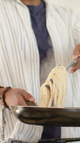 Vertical video: Lifting pasta from pot with tongs, cook in striped top guiding into pan finishing