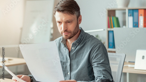 Obraz na plátně Focused Caucasian Male Professional In His 30s Reading Documents At Modern Office Workspace, Sitting At Desk With Laptop And Papers, Engaged In Work