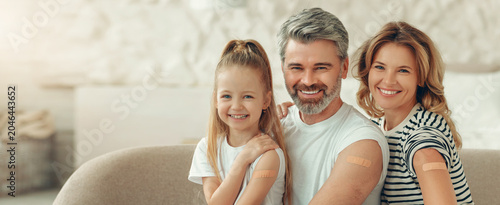 Obraz na plátně Smiling Family Of Three Including Mother, Father And Young Girl Showing Bandages On Arms After Vaccination, Sitting Comfortably Together In Cozy Living Room At Home