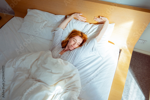 Top view Middle aged Caucasian woman waking up with a smile stretching in bed on a sunny morning. Good morning vibes. Early wake ups routine. Selective focus.
