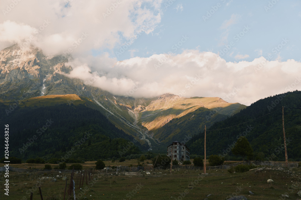 Naklejka premium Quiet mountain valley scene shows a small weathered house in a grassy field beneath dramatic clouds with distant peaks forming a rugged backdrop and a sense of solitude.