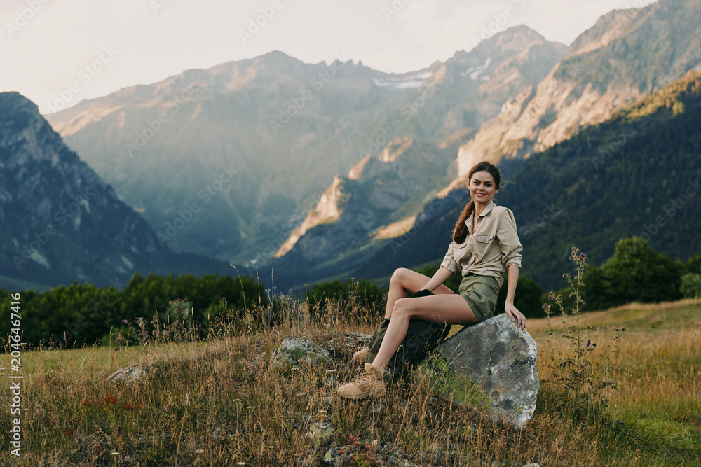Naklejka premium Young woman sits on a rock in a grassy meadow with distant mountains and a clear sky, enjoying a peaceful outdoor moment during a sunny day.