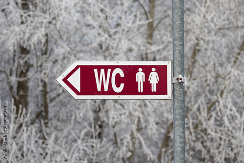 Red WC directional sign with gender icons on a pole against snowy trees