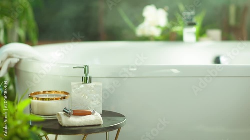 Tranquil bathroom with white bathtub, lush greenery & white flowers. A small table holds a candle, soap dispenser, razor, & towel, creating a luxurious home spa experience for self-care & hygiene.