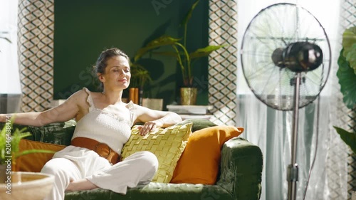 A serene woman on a green velvet sofa, with eyes closed and a gentle smile, enjoys the cooling breeze from a standing fan in a sunlit living room, capturing tranquil heat relief.