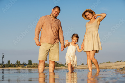 A happy family photo. The family is strolling along the seashore, going slightly into the water. A father, mother, and little girl are relaxing on the beach.